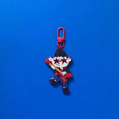 Designerski brelok Rockman Mick z czerwoną gitarą i gwiazdkami, z czerwonym karabińczykiem, na niebieskim tle.